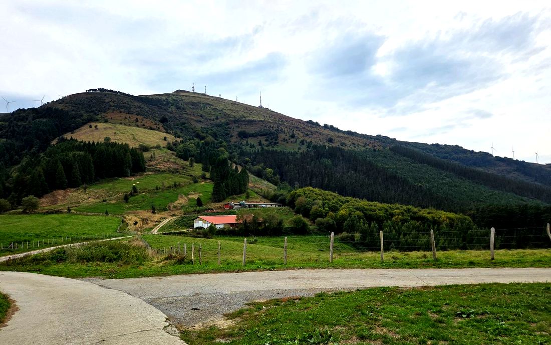 Cruce de pistas de cemento entre prados, con una granja y la ladera del Oiz