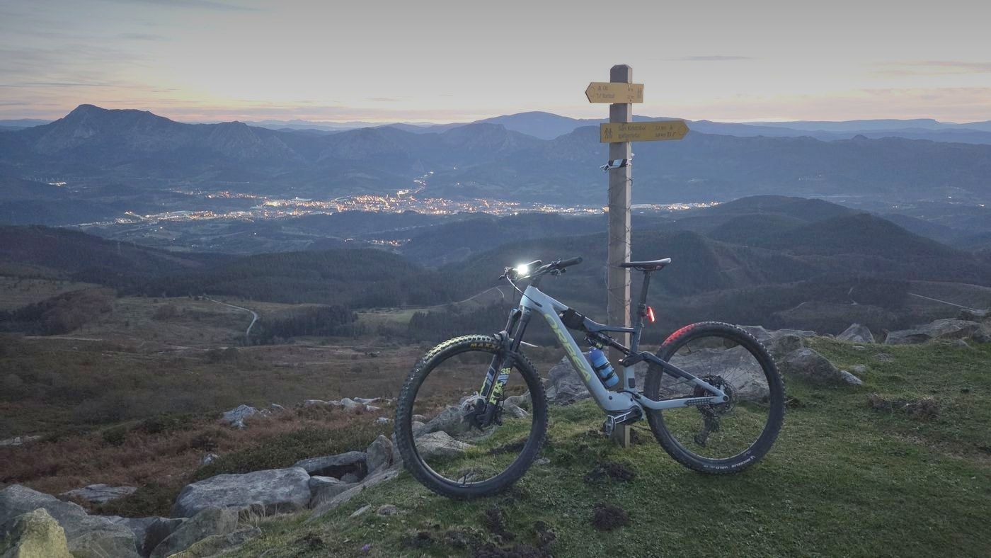 Bicicleta de montaña junto a un poste de señales en el monte, al atardecer, con Mallabia iluminada al fondo