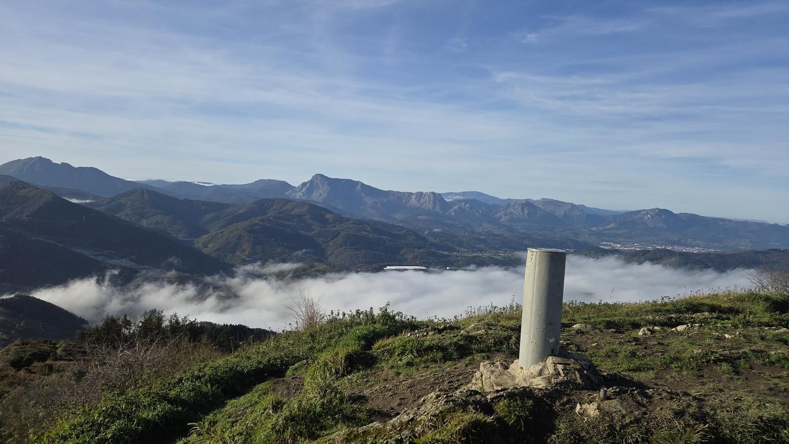 Vértice geodésico en la cumbre del Urko, con nubes bajas y las montañas del entorno al fondo